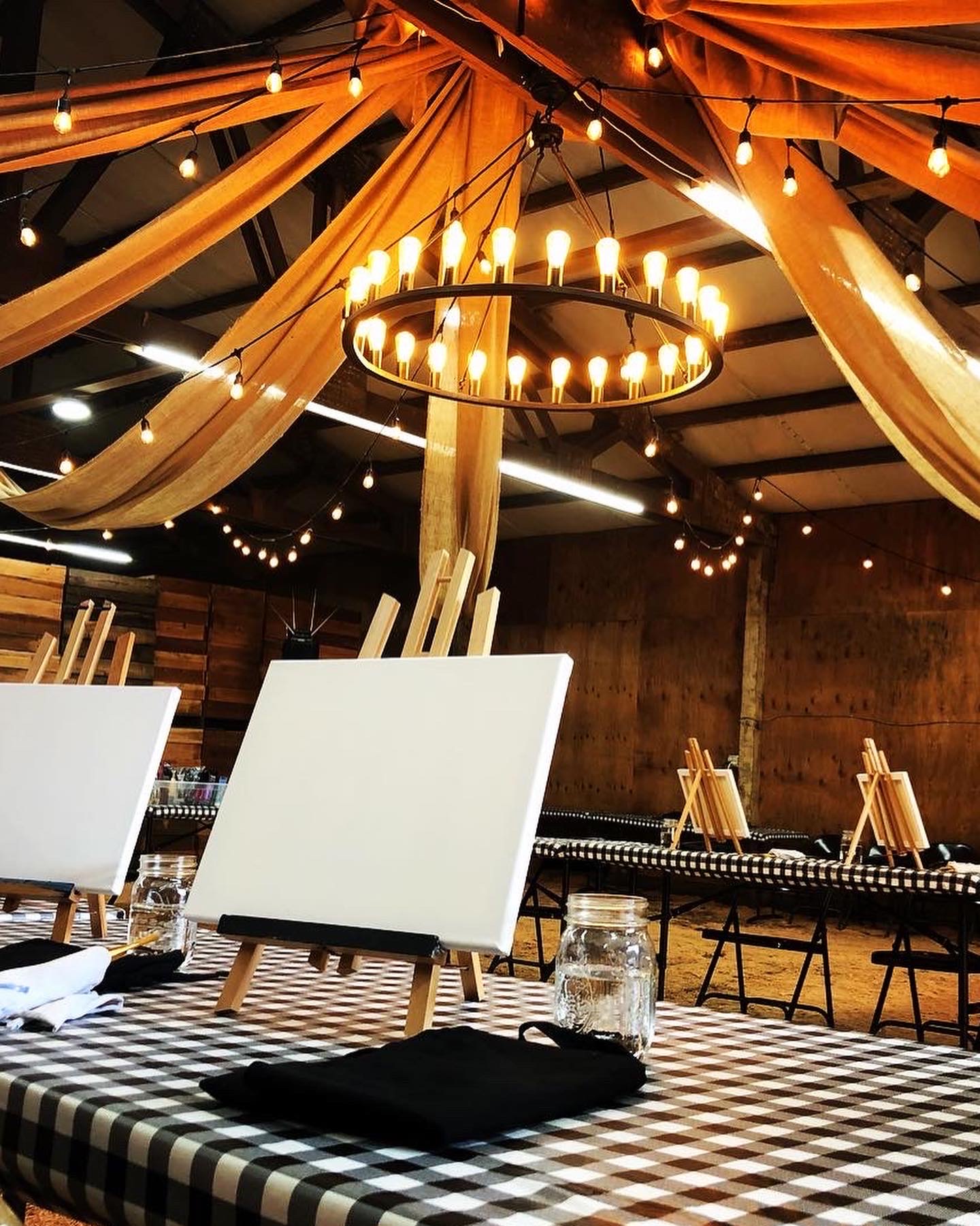 A warm, cozy space with string lights and a chandelier overhead. Tables with checkered cloths hold easels and blank canvases, ready for a painting event.