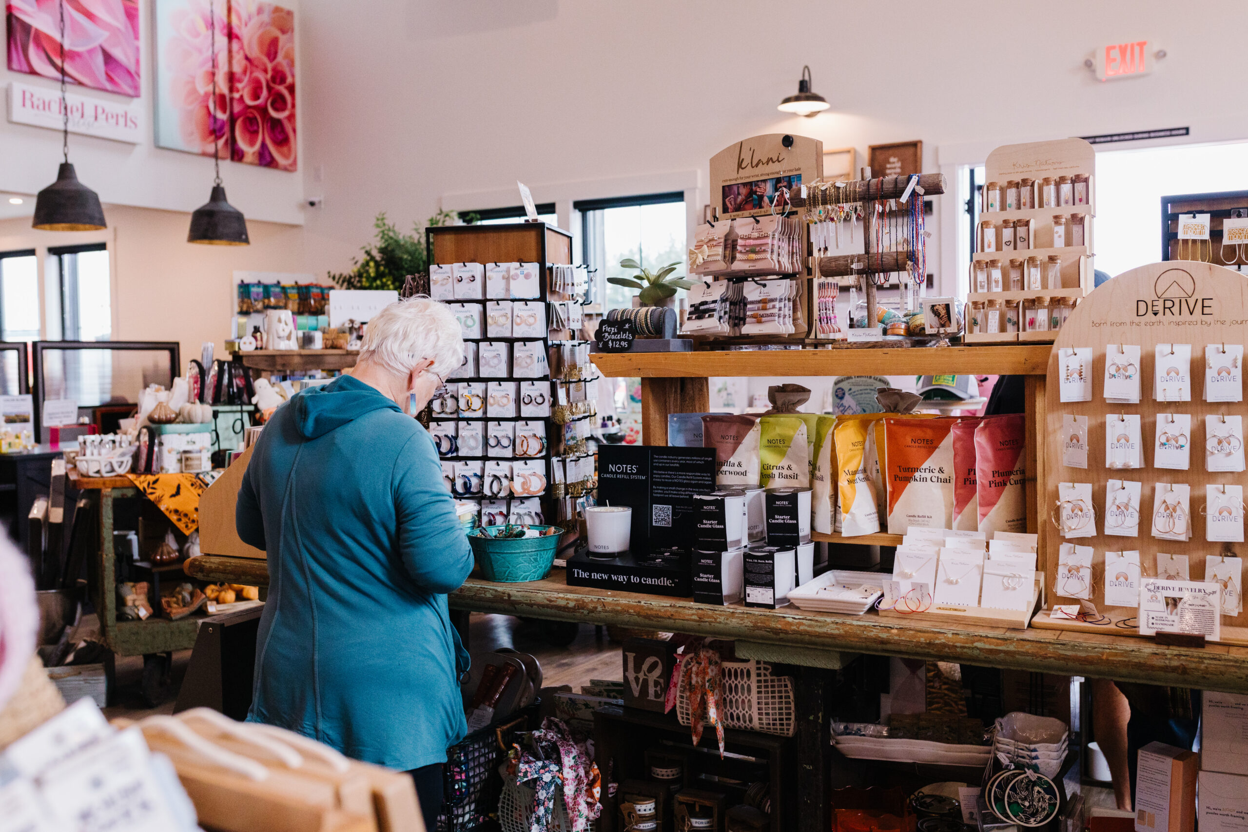 A person with short gray hair shops at a boutique filled with shelves of candles, jewelry, and other gifts. Colorful product packaging is displayed.