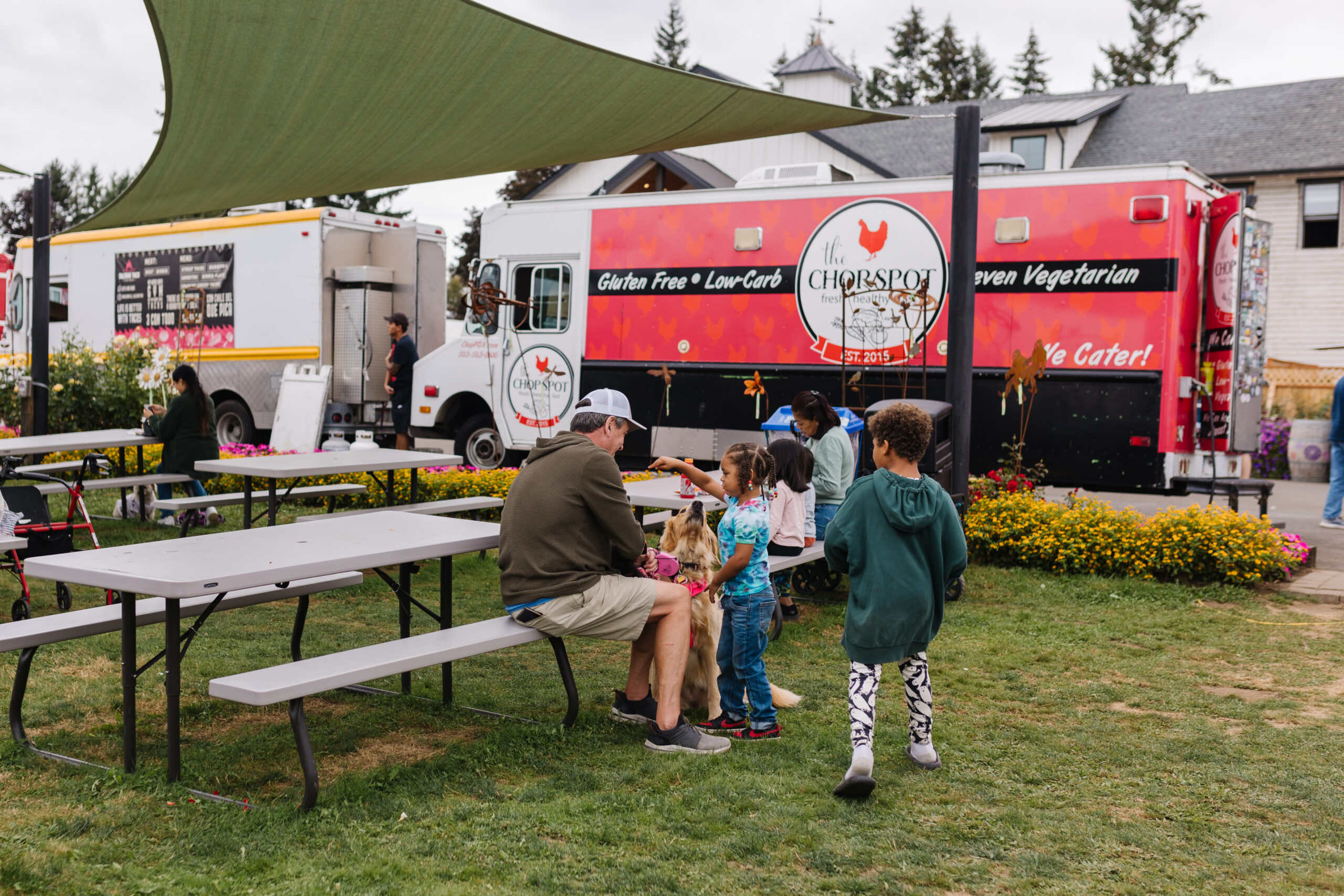 A food truck with the logo "The Chopspot" is parked at the festival. A man and children sit at picnic tables while a dog interacts with the group.