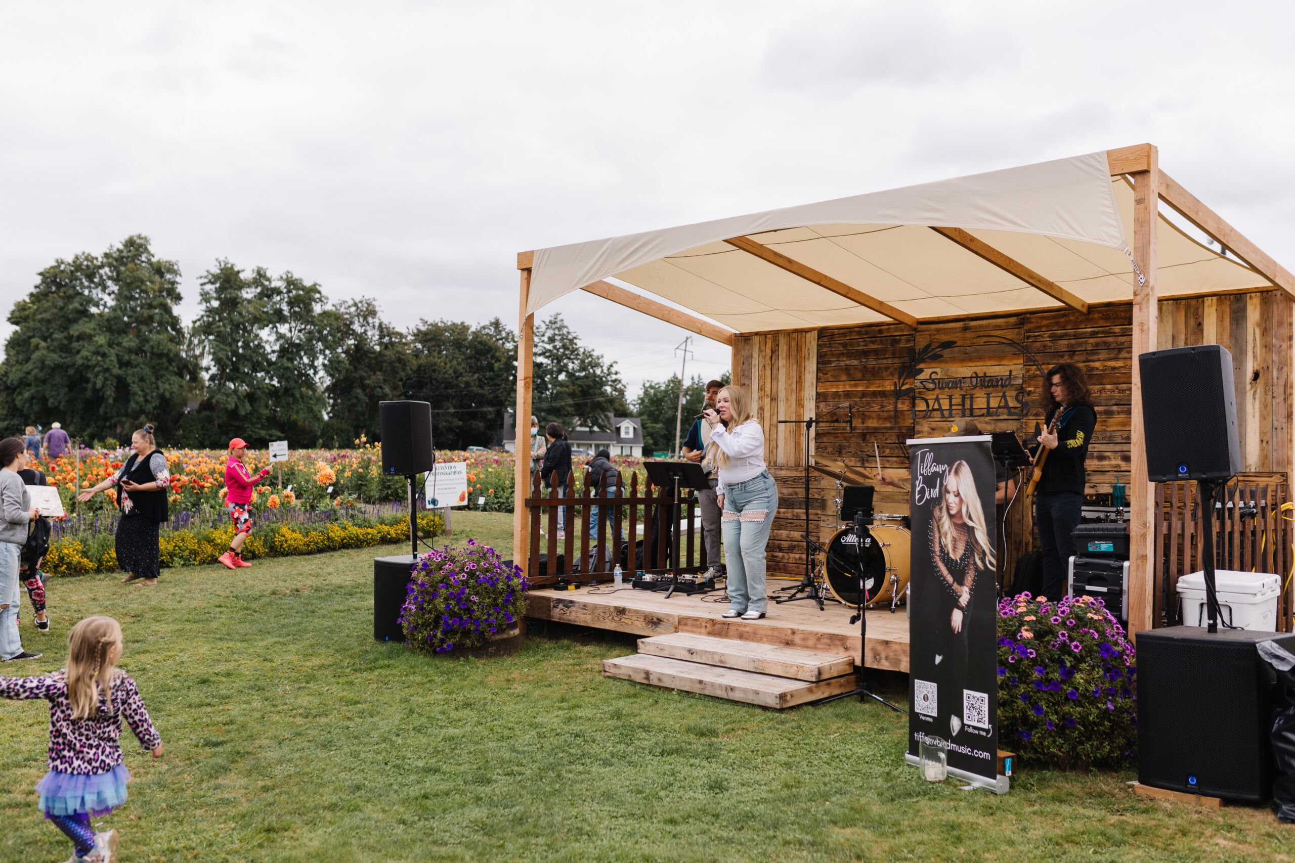 A woman sings on a small wooden stage with a canopy while a child dances in the foreground. Colorful flowers and festivalgoers fill the background.